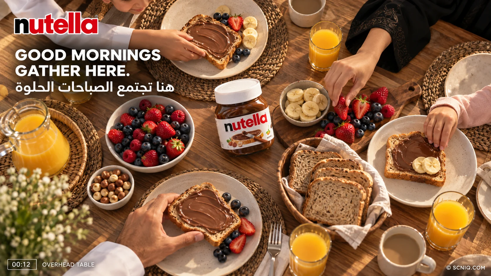 Overhead view of a breakfast table with Nutella toast, berries, sliced banana, orange juice and the jar in the middle as family hands reach in
